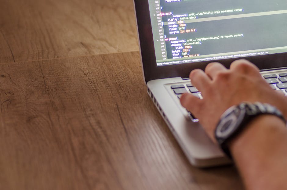 A person's hand is seen typing on a silver laptop with a black keyboard, positioned on a wooden surface. The screen displays lines of code in a text editor, with some commands and syntax visible, sugg
